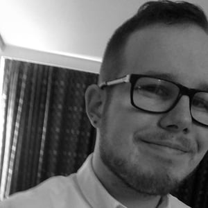 A black and white headshot of James Sedman, a man in his 20s with short, dark hair and short facial hair, wearing a white shirt and glasses and smiling into the camera