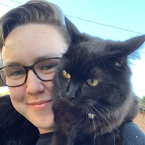 A headshot of Rachael OBrien, a woman with short dark hair and glasses, with her black fluffy cat draped around her shoulders