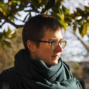 a person with short hair, glasses and wrapped in a green scarf stands below a tree, looking to the side