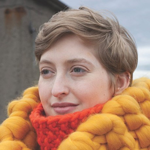 Allie Wheeland a white woman with short blonde hair and grey eyes wearing a bright orange scarf and looking to the side of the camera