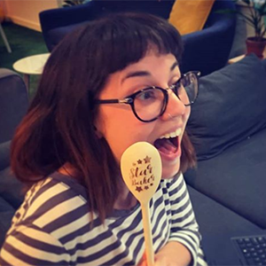 Amy OConnor, a white woman with glasses and dark brown hair, looking delightfully surprised and clutching a Spektrix Bake-Off finalist spoon that she just won.
