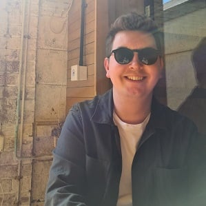 Conor Marum, a white man in his 20s, is enjoying a rare sunny day in Manchester, with sunglasses on, as well as a big grin!