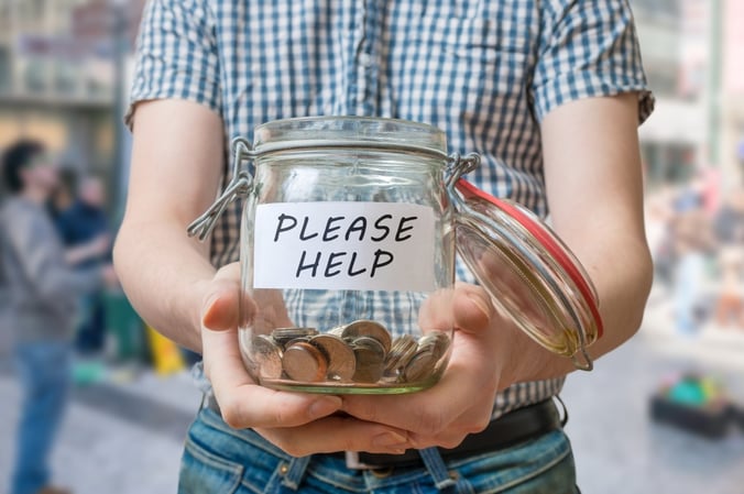 A young figure holds out a glass jar containing coins with a sign saying 'Please Help'
