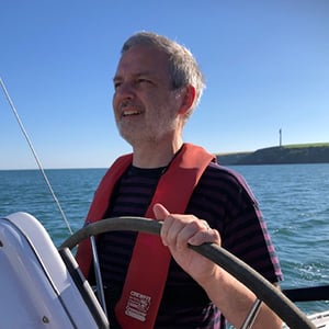 Harry Brett-Jones, a middle-aged white man is wearing a T-shirt and helming a yacht on a summers day. He is looking away from the camera towards the front of the boat. There is blue sky, sea and some land in the background.