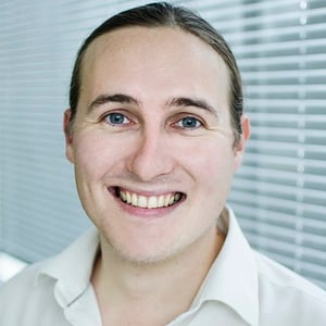 A headshot of Martin Counter, a man in his thirties with long brown hair tied back in a pony tail. He is wearing a white shirt, standing in front of some window blinds and smiling at the camera