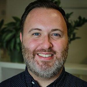 A headshot of Mark Smith, a man with brown hair and a full beard, wearing a button down shirt and smiling into the camera.