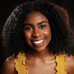 A headshot of Nickasey Freeman, a smiling black women in her early twenties with curly shoulder length black hair wearing a frilly, mustard yellow, tank top.