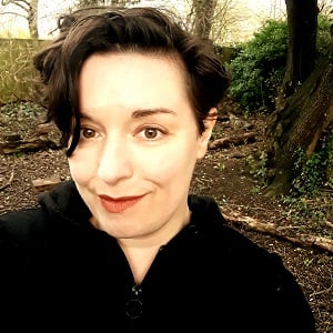 A photo of Melissa Cox, a woman with short, brown wavy hair, wearing a black hoodie, standing in a park with some trees visible in the background.