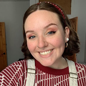 A photo of Caitlin Hudecheck, a white woman in her twenties, with curly brown hair and blue eyes, wearing a red sweater and cream overalls, standing inside of an apartment with white walls.