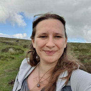 A selfie of Amy Brown, a white woman in with long light brown hair, looking windswept smiling into the camera on a hillside during a hike.