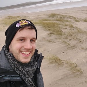 A selfie of Darko with a toothy smile, wearing a grey winter coat, grey thick scarf and a black Lakers beanie with a bit of brown  fringe sticking out. He is standing on a Scottish, sandy beach on a grey day with foamy waves heading for shore.