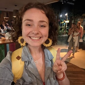 A selfie of Ruth Brown, a women with brown curly shoulder length hair, wearing a jean jump suit, yellow backpack and big sunflower knitted earrings, smiling into the camera.