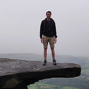 Chris Taylor, a white man in shorts and a rain coat, poses for a photo when standing on a rock ledge in the Peak District on a grey day.