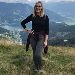 Dani Gambier, a white woman with blonde hair and glasses, stands with a hand on hip, with a backdrop of a lake and mountains in the Alps.