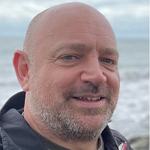 David Marshalsea, a man in his forties with shaved hair & stubble , stands in front of the sea at Porlock Wier.