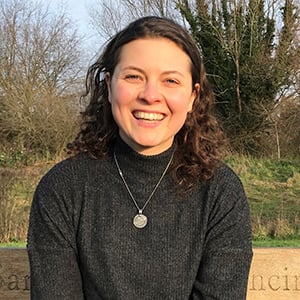 Georgie Pepper, a woman with brown curly hair, sitting on a bench in a park and smiling to the camera