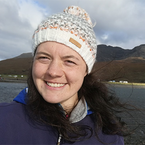 Hannah Wisdish, a woman with brown wavy hair, wearing a woolly hat and smiling, stands in front of a lake with hills in the background.