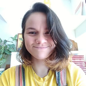 Headshot of Lowri Jones with yellow top and orange dungarees, background is a colourful bookcase.