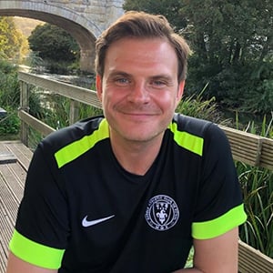 James Bush, a man in his late 30s wearing his 5-a-side football kit sits beside a river