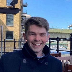 Jamie Wright, a young white man with short brown hair, sits smiling next to the River Thames.