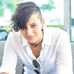 A picture of a non-binary person, Dex Manson, with short, black hair, wearing a white long sleeved shirt and blue pants smiling into the camera.