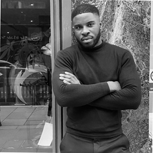 Oliver a man in a black turtle neck leans against a building with crossed arms