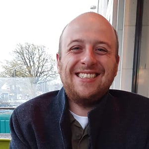 Ollie Freeman, a man in his twenties with a beard, sitting outside wearing a grey coat, and smiling into the camera.