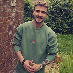 Raice Bassett, with short hair wearing a green jumper, stands in an outdoor environment