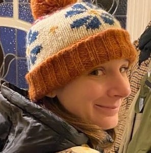 Sarah Frost, a woman wearing an orange bobble hat, is looking over her shoulder and smiling