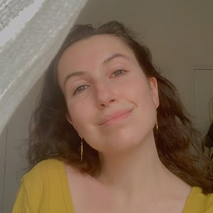 Daria Kerschenbaum, a white woman with dark hair, smiles into the camera while peaking out from a lace curtain, wearing a yellow shirt.