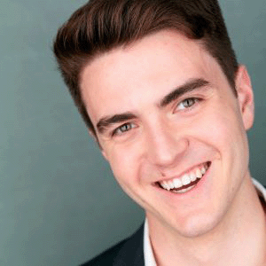 a headshot of Dean, a man with short light brown hair, wearing a suit jacket over a white shirt, smiling into the camera