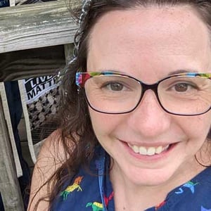 Laurice Grae is a middle-aged white woman with curly dark hair and glasses, smiling outdoors